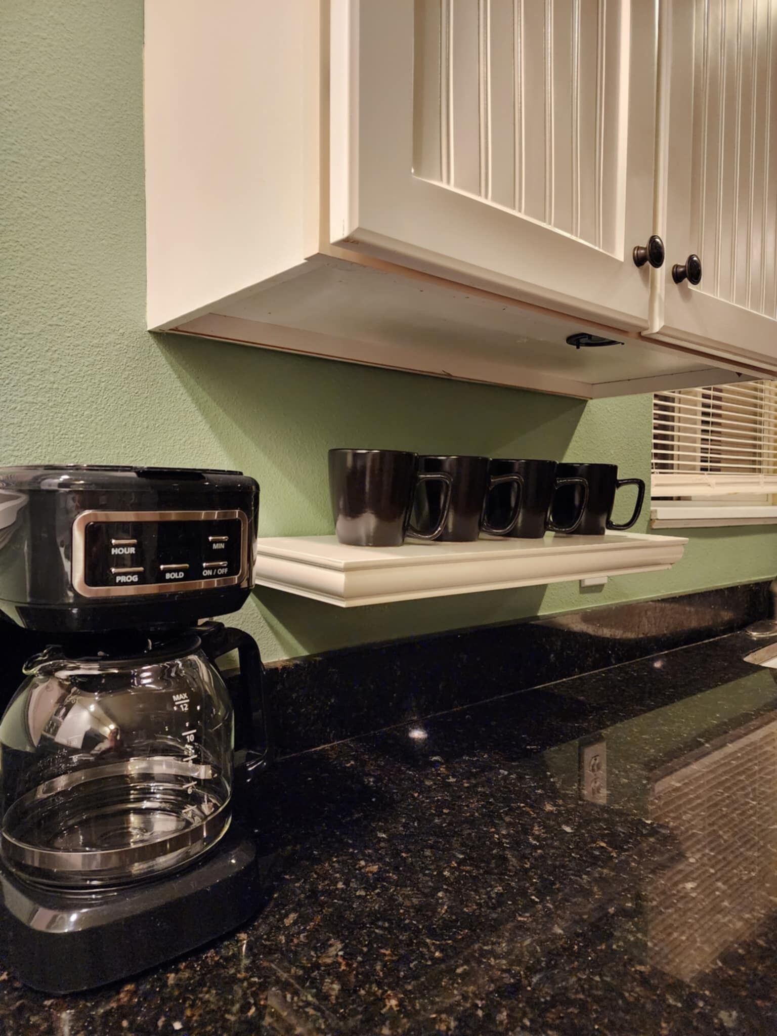 Une machine à café est posée sur un plan de travail dans la cuisine, et une étagère au-dessus contient plusieurs tasses noires.