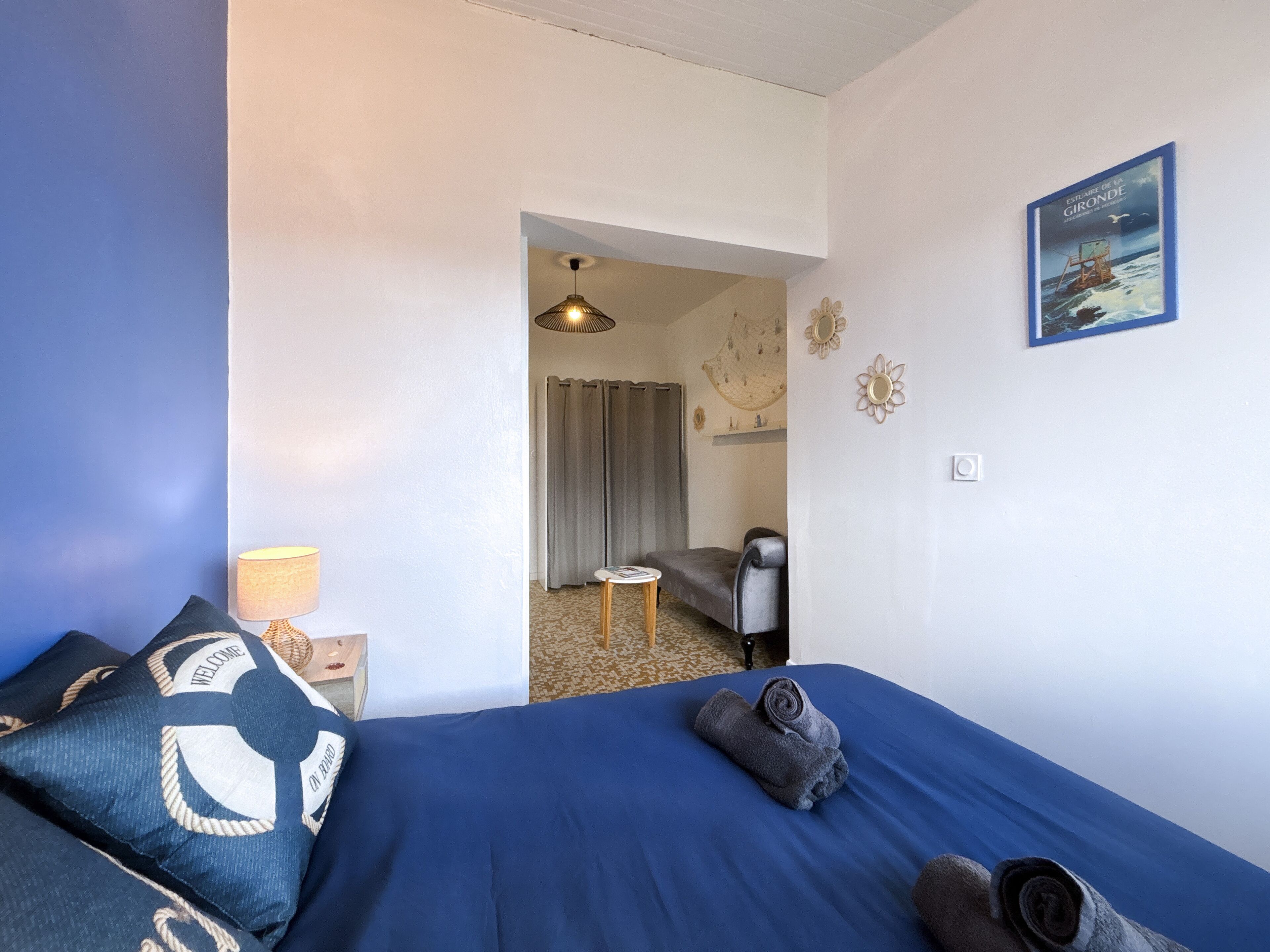 A hotel room with a bed, a small table with a lamp, a chair, a door leading to another room, and a framed picture on the wall.