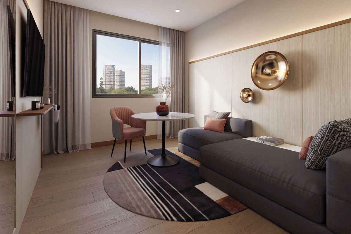 Ein modernes Hotelzimmer mit einer Couch, einem kleinen runden Tisch, einem Fernseher und Blick auf Gebäude durch große Fenster.