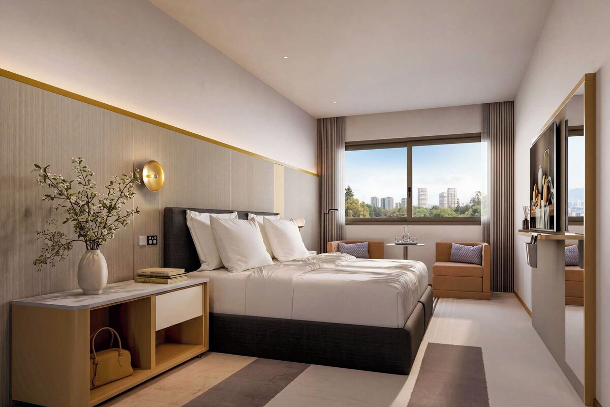 Ein modernes Hotelzimmer mit einem großen Bett, Nachttischen, einem Schreibtisch, einem Sessel und einem Fernseher. Ein Fenster bietet Blick auf die Stadt.