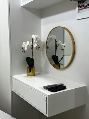 Ein Badezimmer mit weißem Waschtisch, einer goldenen Vase mit weißen Orchideen, einem runden Spiegel mit goldener Rahmung und einer schwarzen Fernbedienung auf dem Waschtisch.