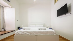 Een opgemaakt bed met witte lakens en handdoeken, een flatscreen televisie aan de muur en een houten vloer.