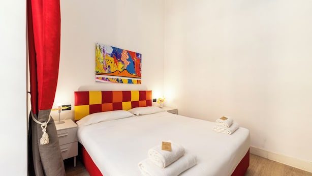 A hotel room with a bed, a nightstand, a lamp, a painting, and a red curtain. - Casa di Rosy (Naples)