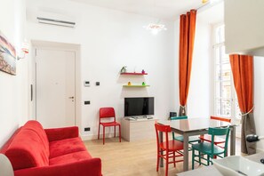 A compact living space with a red sofa, a dining table with chairs, a TV, and a window with curtains. - Casa di Rosy (Naples)