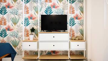 Una habitación con un televisor sobre un mueble bajo, un pequeño armario blanco con cajones, una planta en maceta y una silla azul.