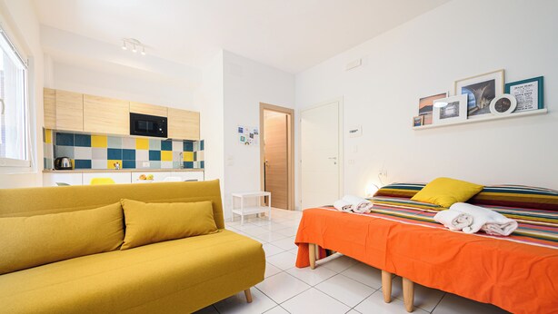 A modern hotel room with a yellow sofa, an orange bed with striped bedding, a white wall with framed pictures, and a kitchen area with a microwave and tiled backsplash. - L'Aurora Apartments (Naples)