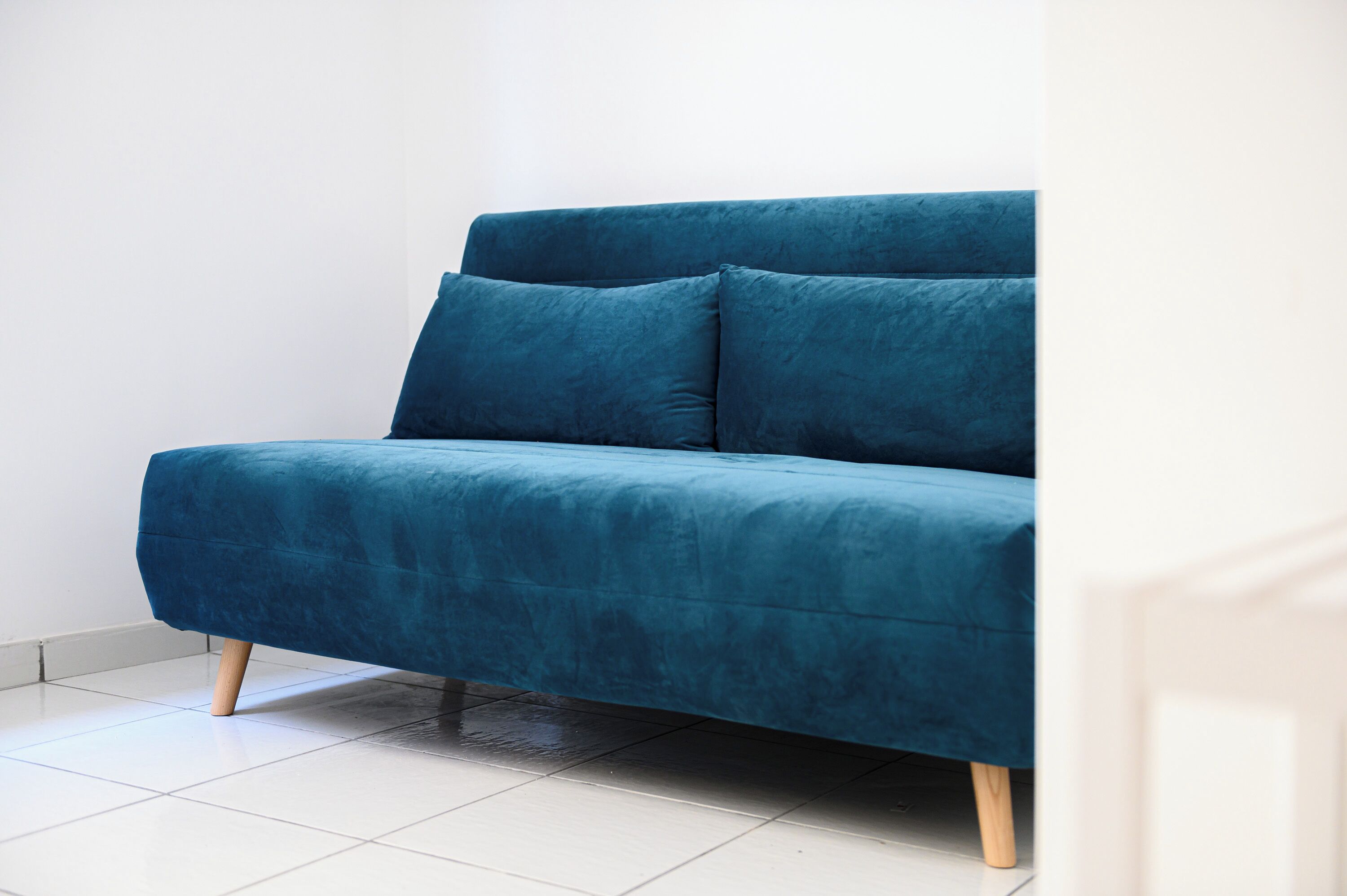 Un sofá azul con patas de madera en una habitación con paredes blancas y suelo de baldosas.