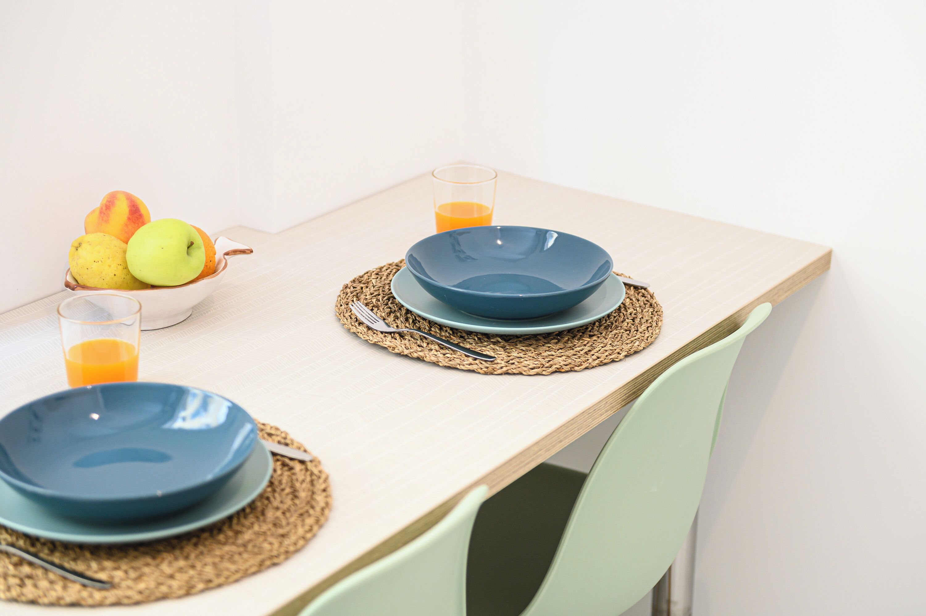 A dining table with two blue plates, a glass of orange juice, and a bowl of fruit.