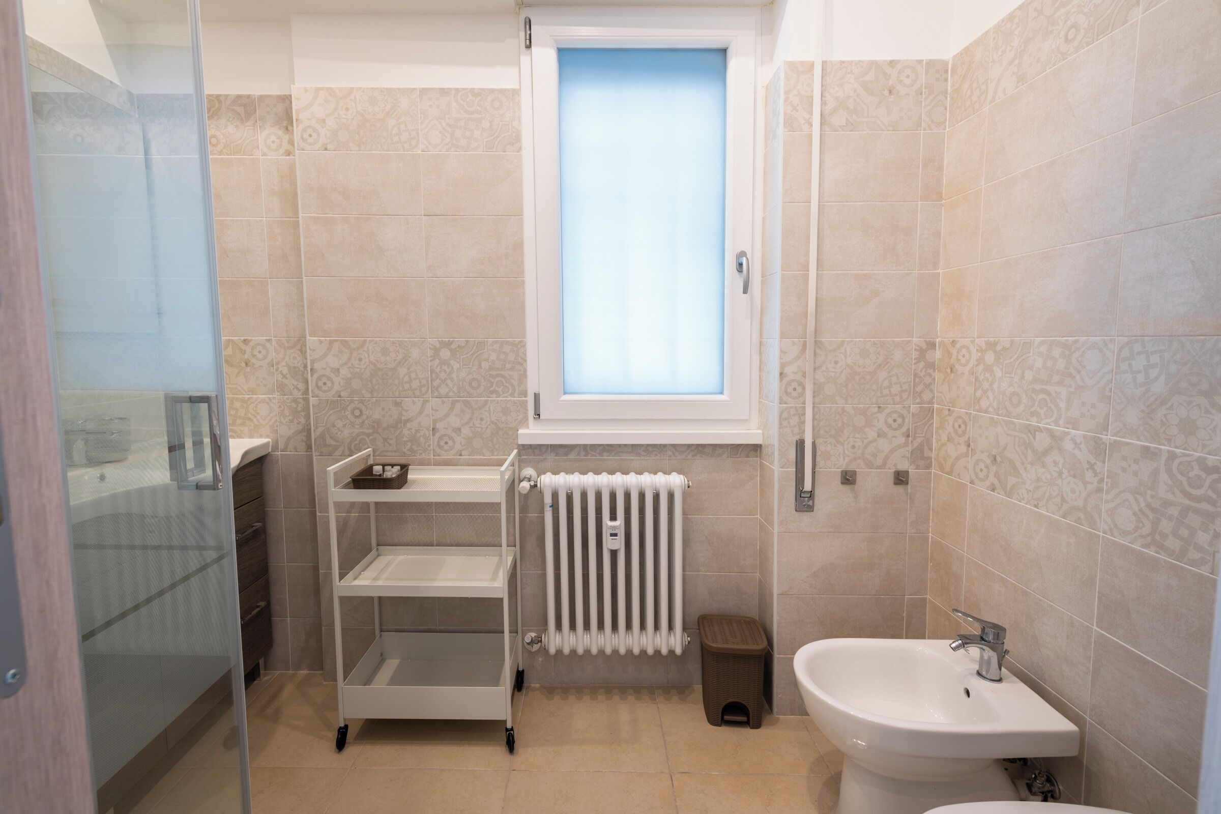 Een badkamer met een glazen douchecabine, een witte radiator, een wastafel en een raam met zonwering.