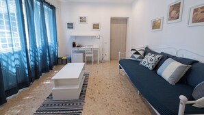 Una sala moderna con sofá, mesa de centro, escritorio y cama. Hay cuadros enmarcados en la pared y una ventana con cortinas.