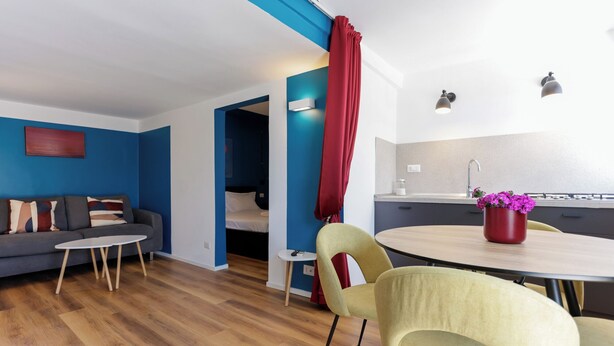 A modern living room with a sofa, a small round table, and two chairs. There is a kitchen area with a sink and a red curtain. - Via Galiani 20 (Naples)