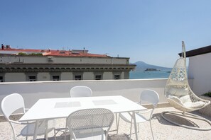 A terrace with a white table and chairs, a hanging chair, and a view of the sea and buildings. - Via Galiani 20 (Naples)
