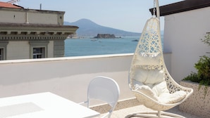 A white hanging chair with a cushion, a white table, and a view of the sea and a mountain in the background. - Via Galiani 20 (Naples)