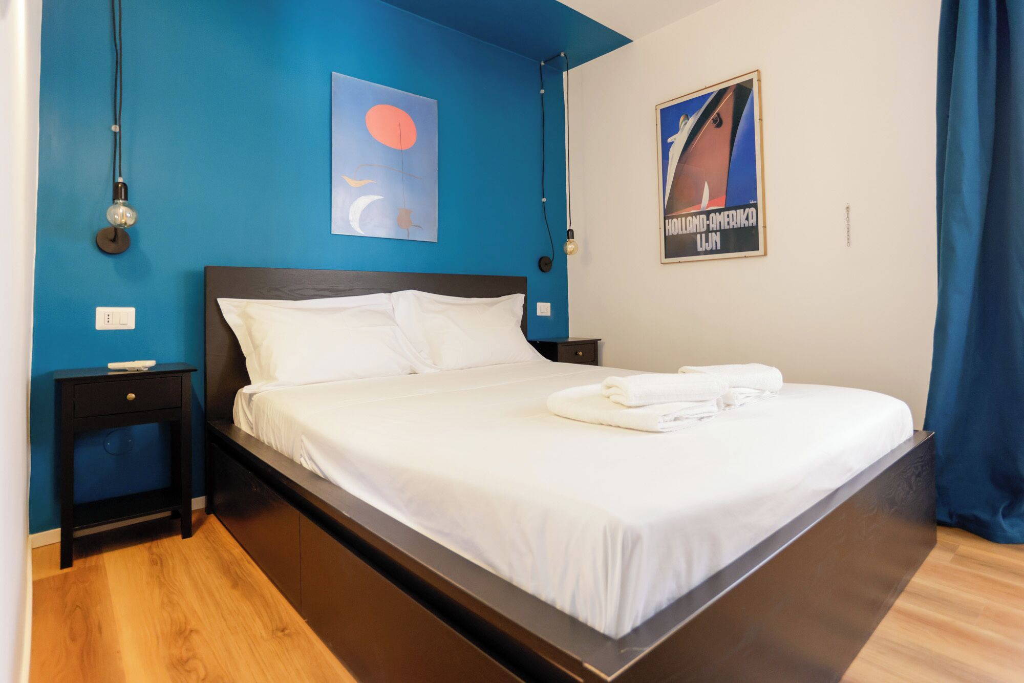 A hotel room with a bed, a nightstand, a wall clock, and a poster.