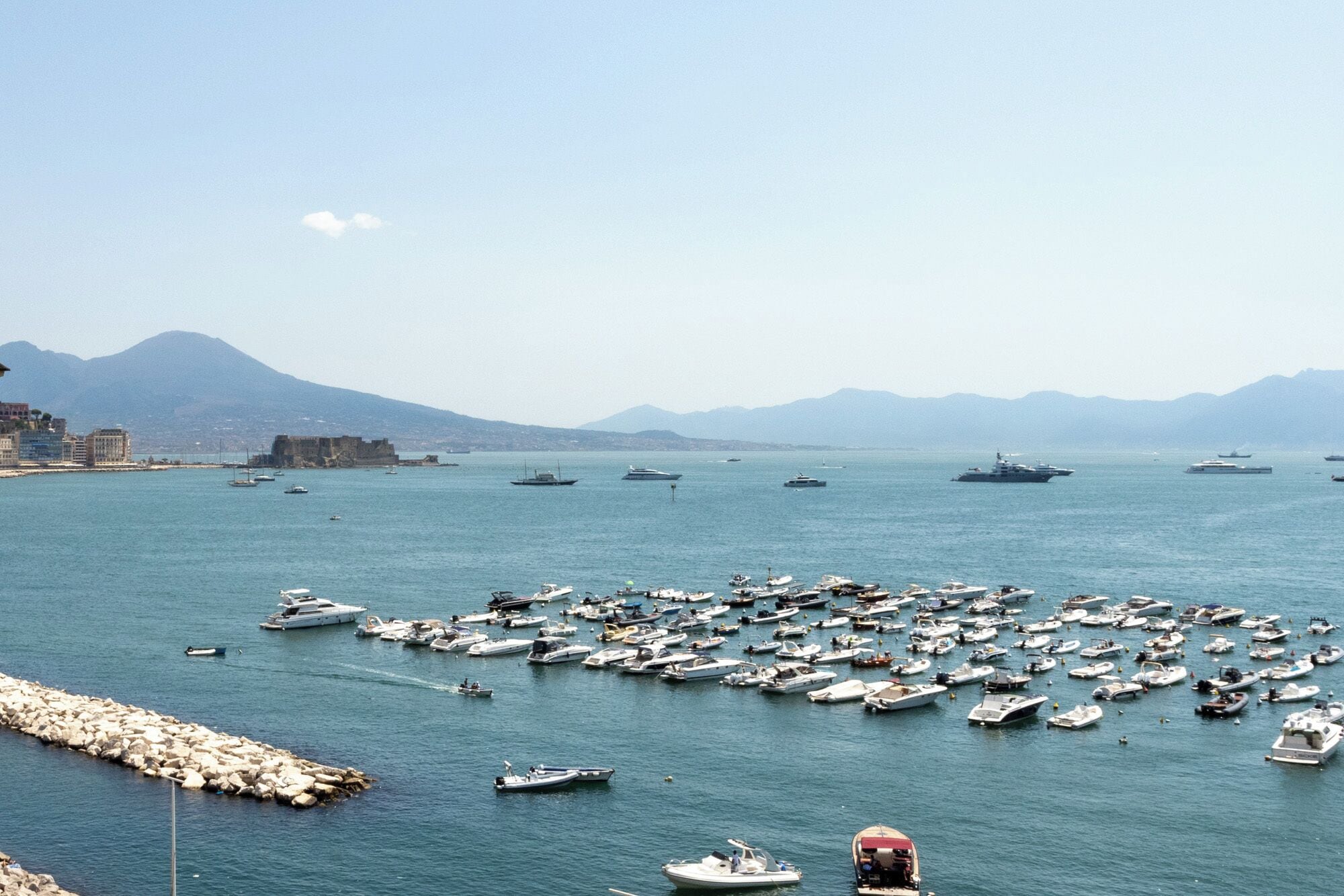A marina with numerous boats docked, a rocky breakwater, and distant mountains.