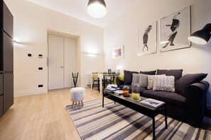 Ein modernes Wohnzimmer mit einem dunklen Sofa, einem Couchtisch und einem Streifenmuster-Teppich. Es gibt einen Essbereich mit einem Glastisch und Stühlen, sowie eine Küchenzeile mit Kühlschrank und Schränken.
