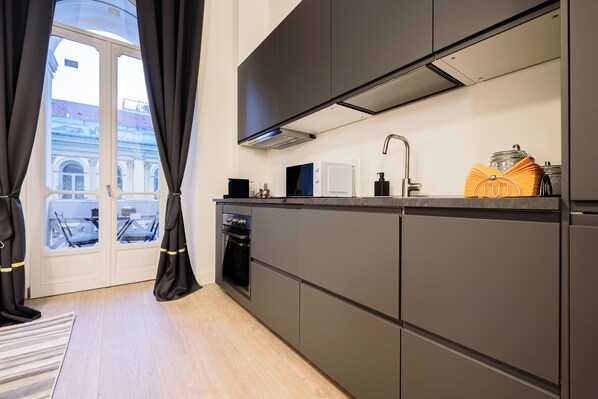 A modern kitchen with dark cabinetry, a built-in oven, and a window with curtains. - Apt503a (Naples)