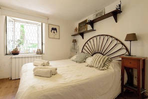 A bedroom with a bed, a window, a radiator, a shelf with decorative items, and a bedside table with a lamp. - Giardino Segreto - Via Foria 216 (Naples)