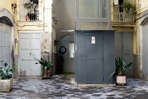 A narrow alleyway with a grey utility box, potted plants, and a metal gate. - Giardino Segreto - Via Foria 216 (Naples)