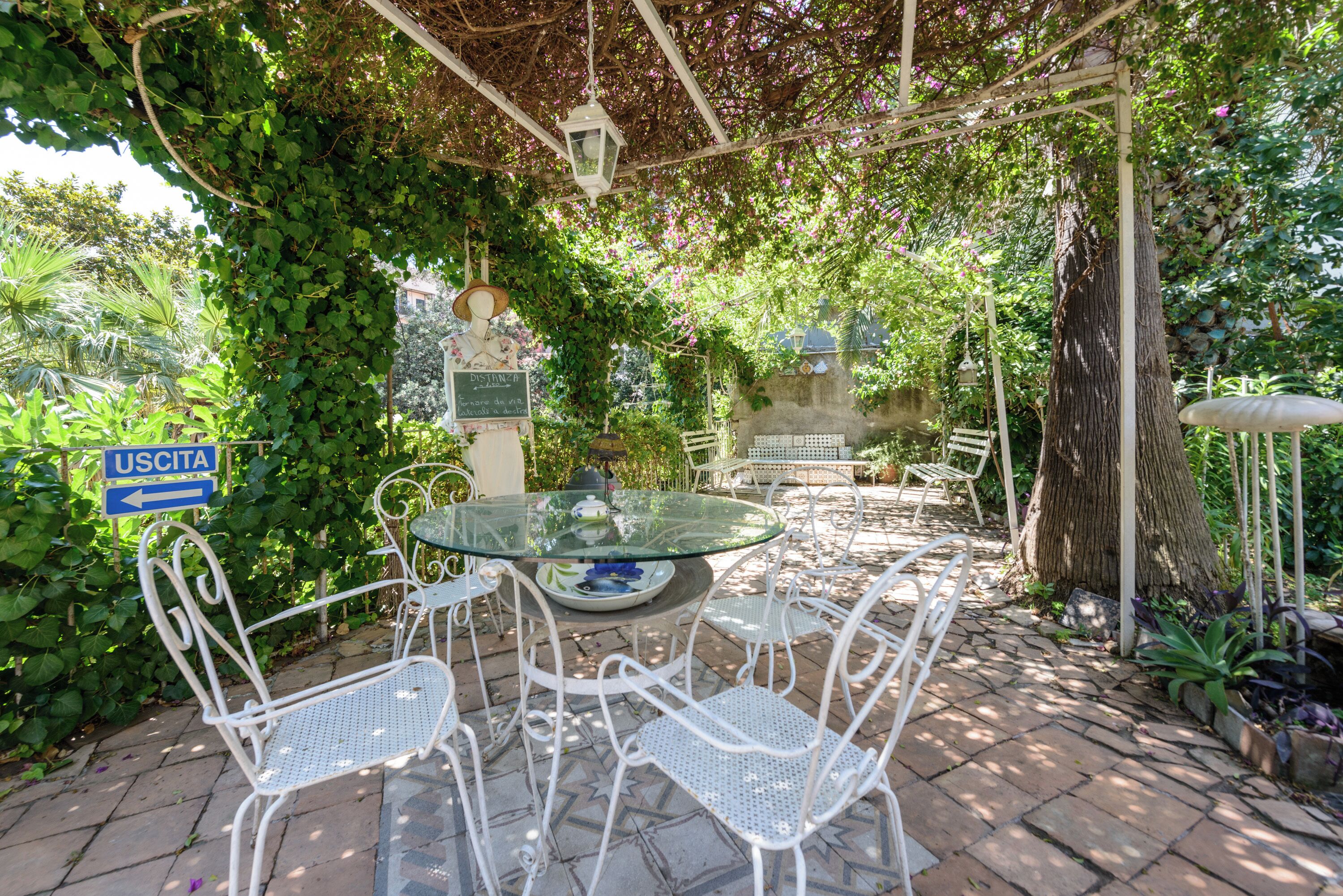Eine Terrassengruppe im Freien mit einem Glastisch und weißen Metallstühlen, umgeben von üppigem Grün und einem Pergola.