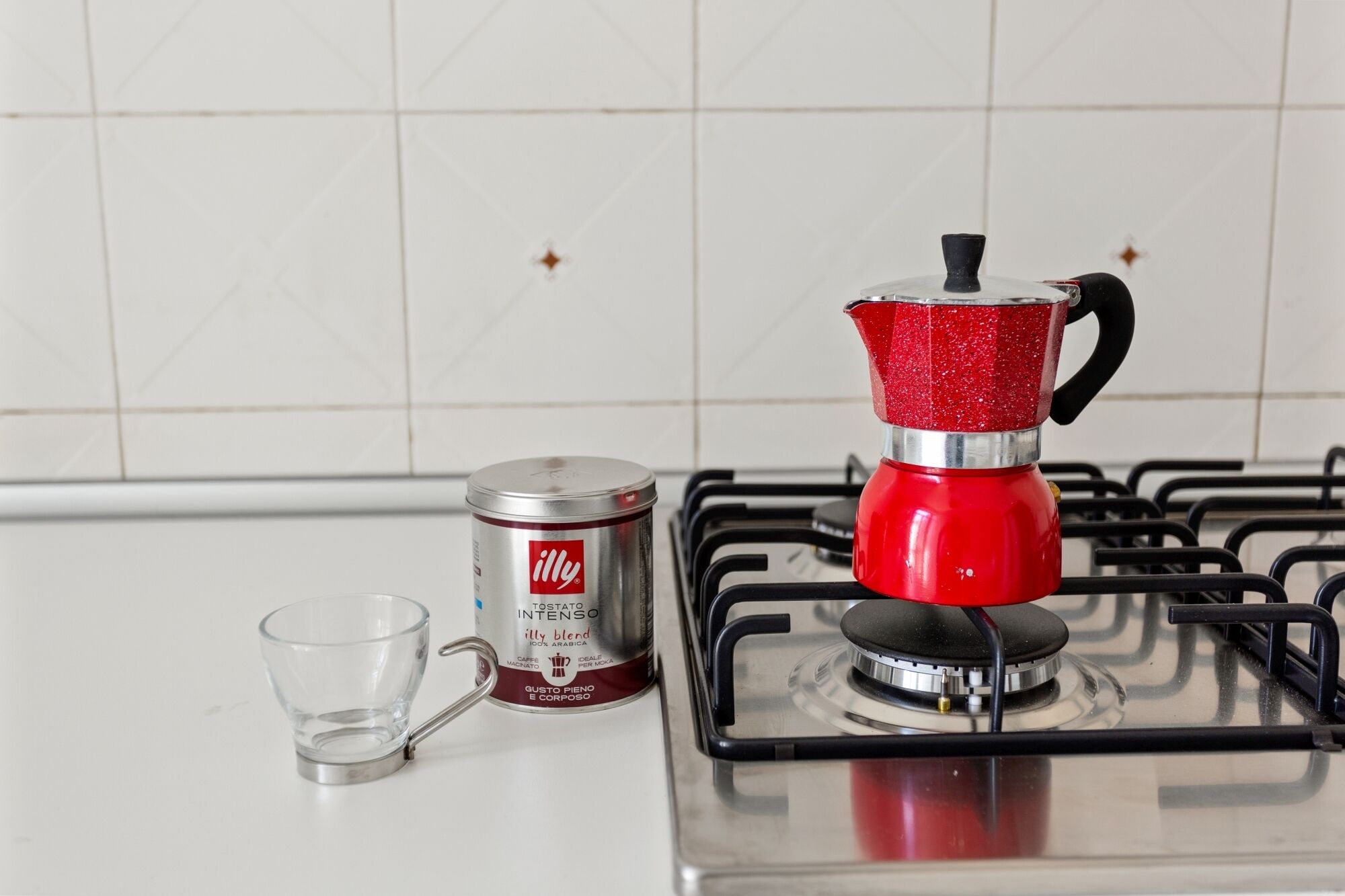 A kitchen with a gas stove, a red coffee maker, a glass cup, and a canister of coffee.