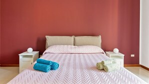Una cama bien tendida con un edredón rosa y dos almohadas, dos mesitas de noche blancas con lámparas y dos toallas enrolladas sobre la cama.