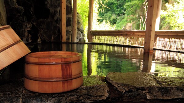A wooden bath tub in a stone-walled room with a bamboo fence and greenery outside.