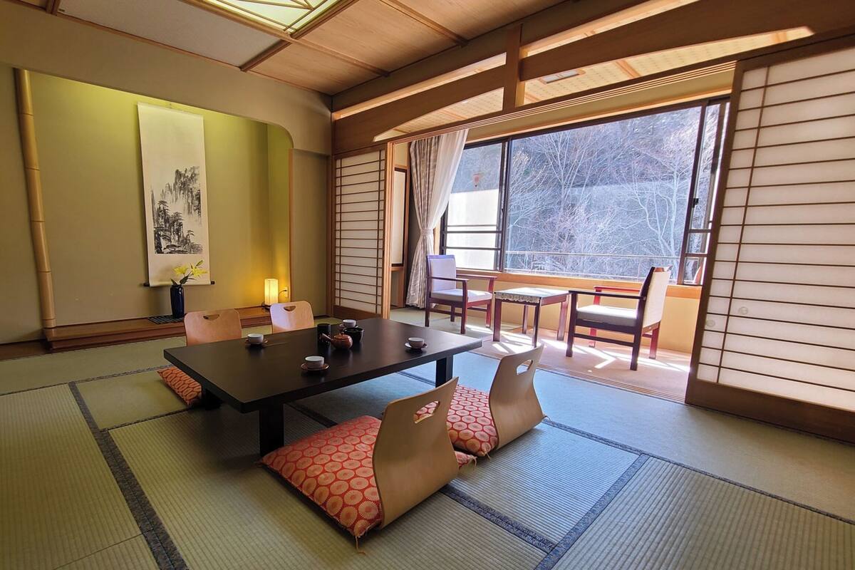 Um quarto japonês tradicional com tatames, uma mesinha com chá, portas deslizantes e vista para árvores lá fora.