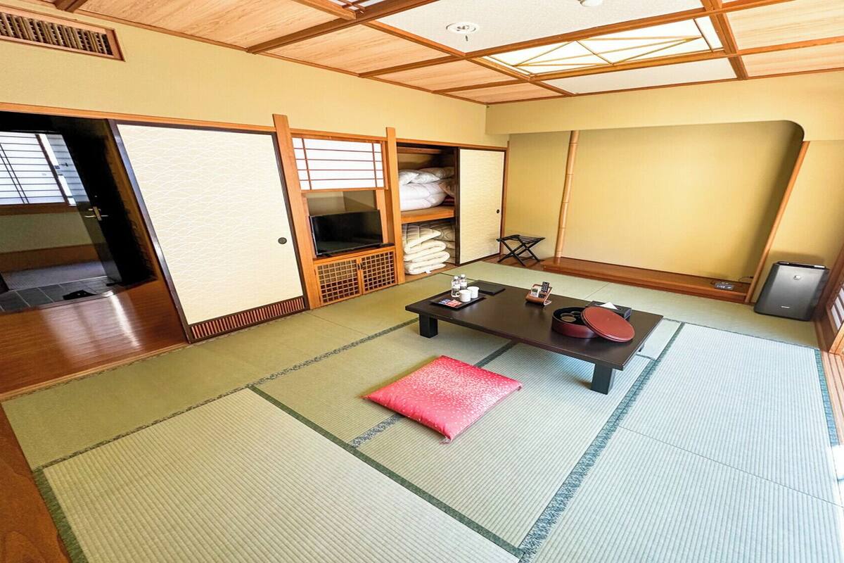 Quarto japonês tradicional com tatames, uma mesa baixa com almofada vermelha, uma TV e um guarda-roupa com portas de correr.