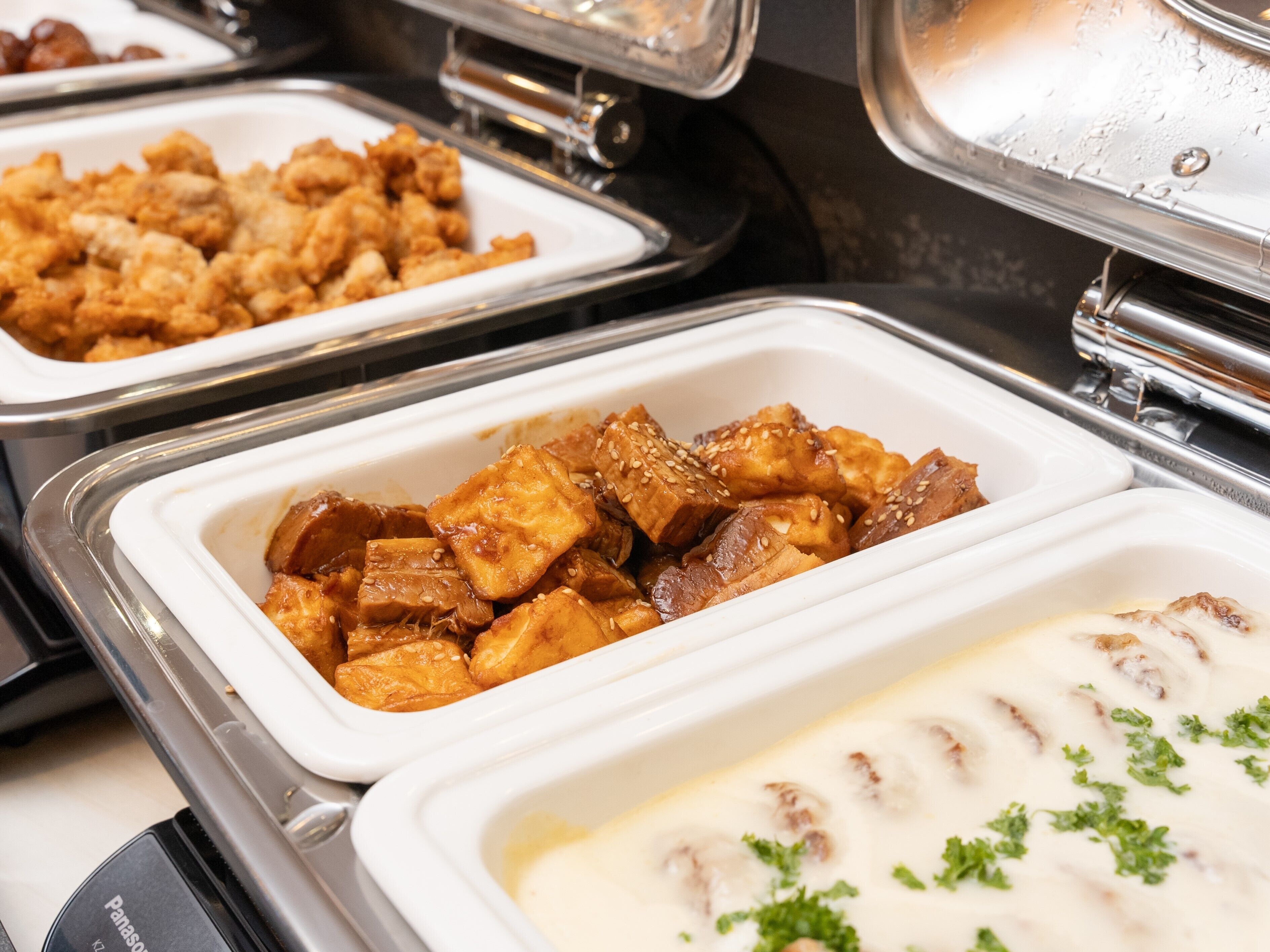 Un buffet proposant divers plats présentés dans des plateaux blancs, notamment ce qui ressemble à du poulet frit et un plat crémeux garni d’herbes.