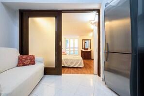 A compact room with a white sofa, a red floral pillow, a bed with a white bedspread, and a modern black refrigerator. - Via Avvocata 25 (Naples)