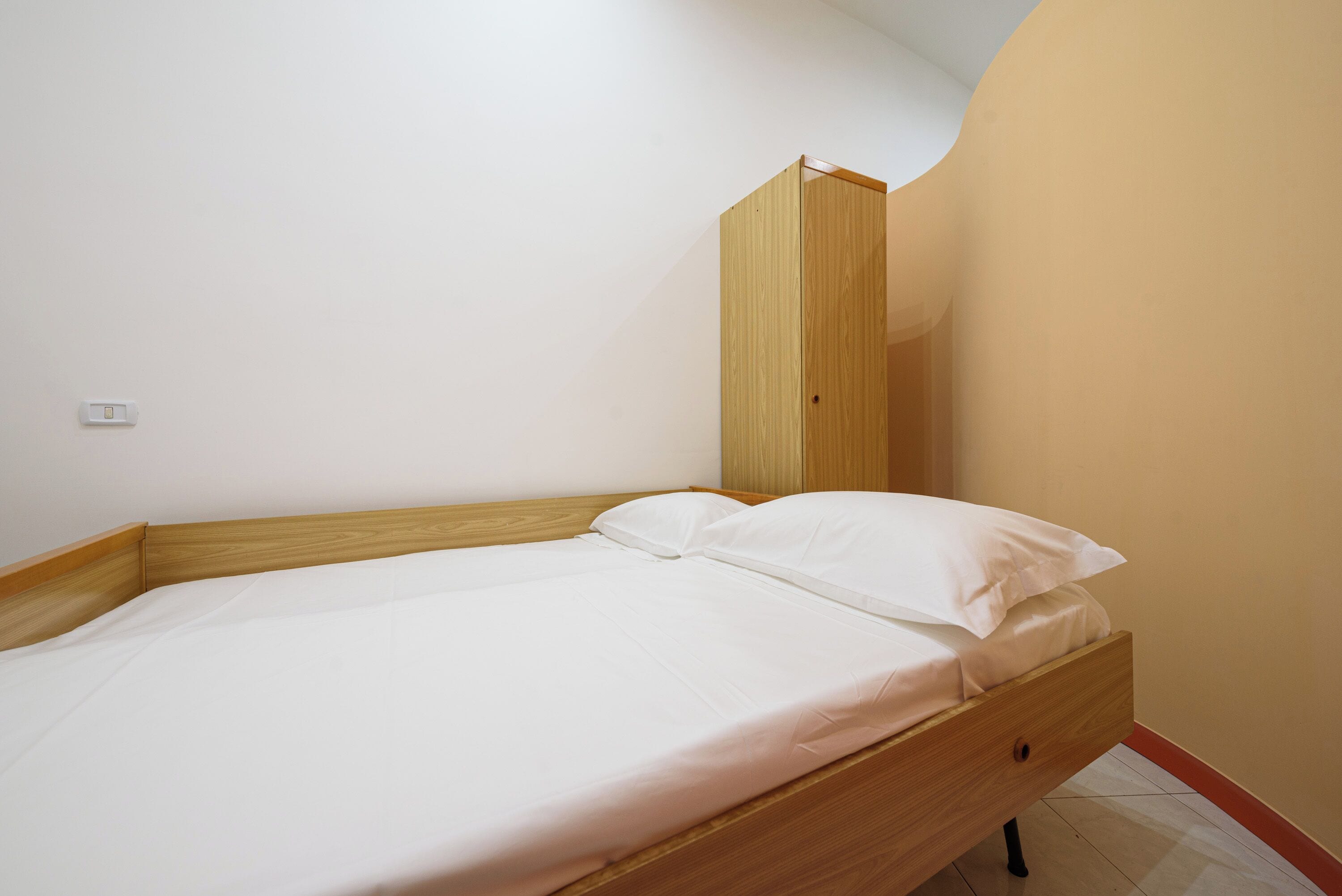 A single bed with a wooden frame and white bedding in a room with a curved wall and a wooden wardrobe.