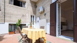 An outdoor patio with a round table covered by a yellow tablecloth, surrounded by wooden chairs. There are potted plants and a tiled floor. - Viale Calascione 7 (Naples)