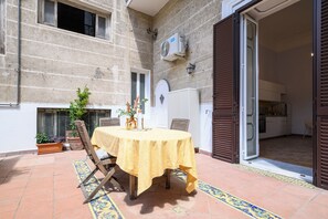 Une terrasse extérieure avec une table ronde recouverte d’une nappe jaune, entourée de chaises en bois. On y trouve également des plantes en pot et un sol carrelé.