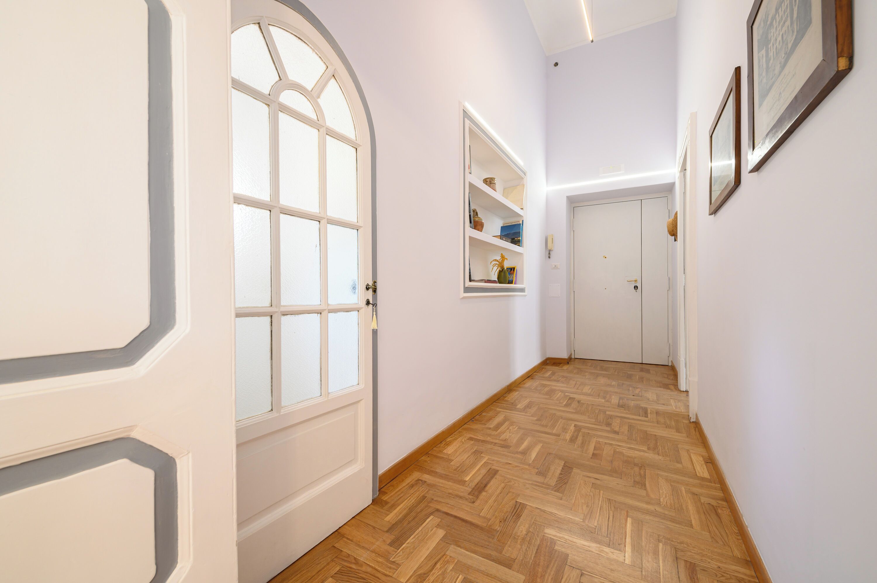 Un pasillo con suelo de madera en dibujo de espiga, paredes blancas y una puerta blanca con panel de vidrio.