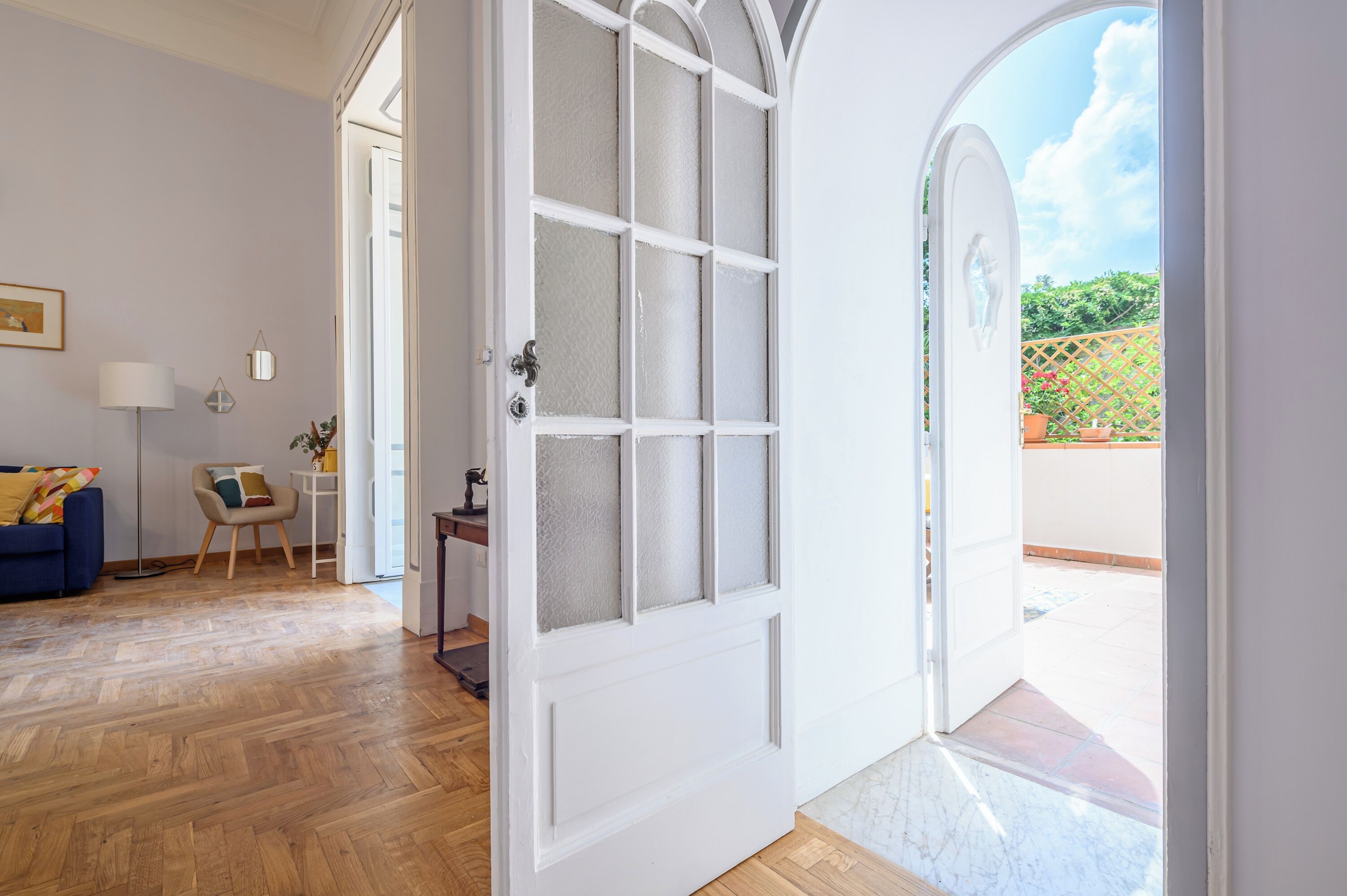 A room with a wooden floor, a white door with glass panels, a blue sofa, a small table with a lamp, and a view of a garden through an arched doorway.