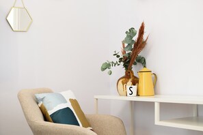 Un fauteuil beige avec un coussin, une table d’appoint blanche ornée d’un vase contenant des plantes vertes et d’un pot jaune, et une lettre « S » posée sur la table.