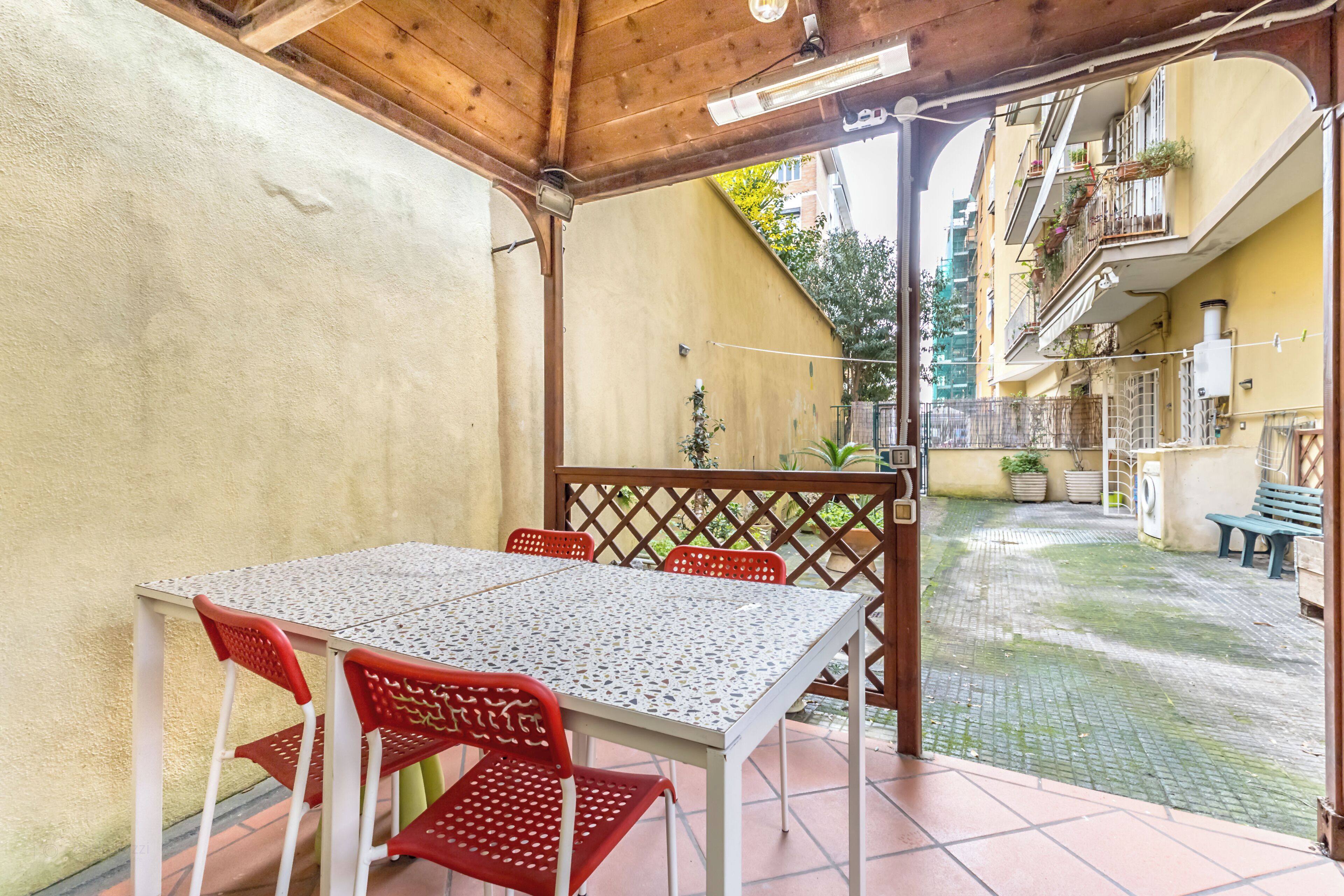 Ein überdachter Patio-Bereich mit Tisch und Stühlen, einer Holzbalkendecke und Blick auf einen Innenhof mit Gebäuden und Pflanzen.