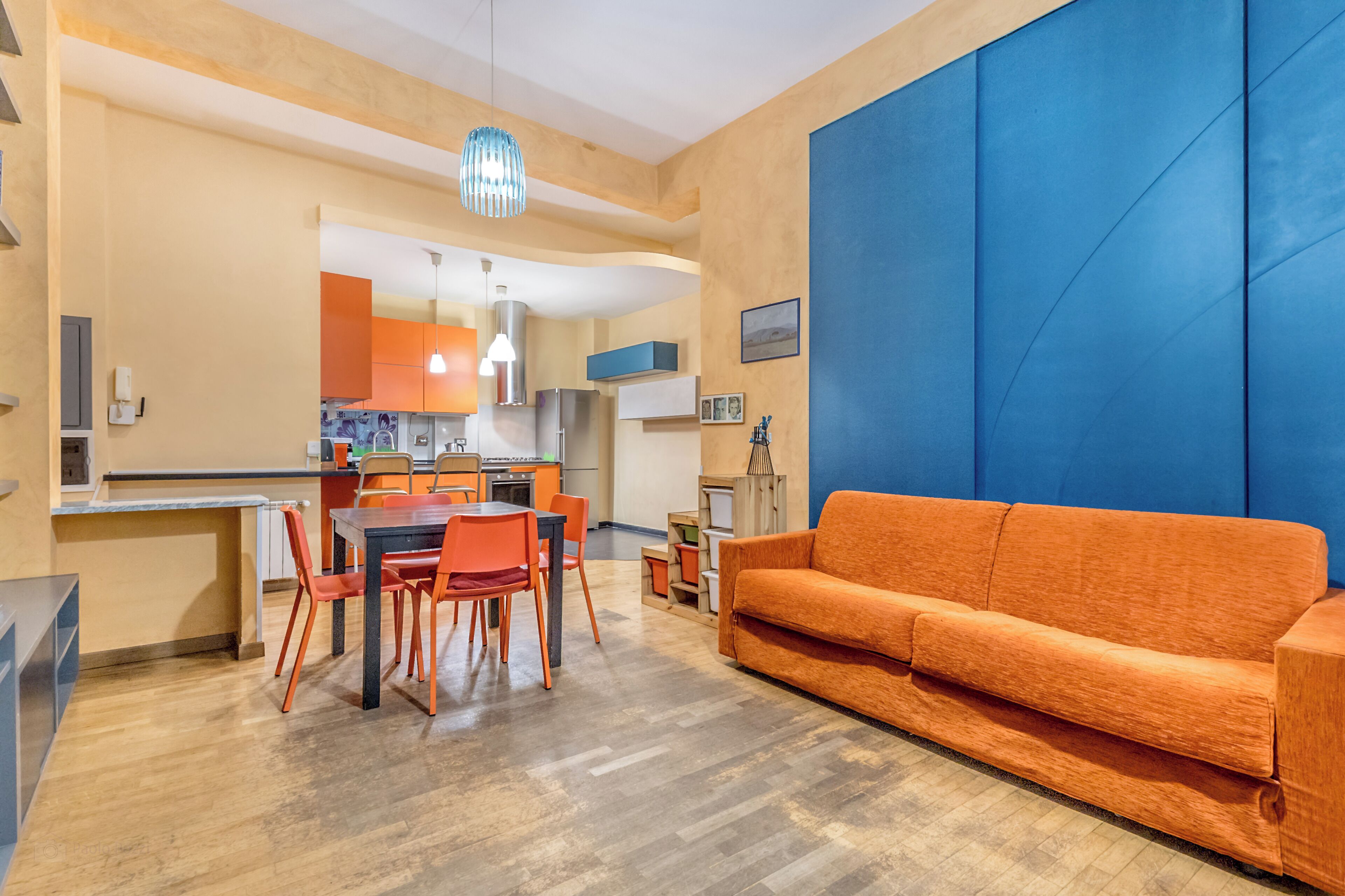 A modern living room with a sofa and dining area, kitchen in the background, and a blue accent wall.