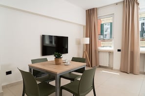 A modern hotel room with a wooden dining table, green chairs, a flat-screen TV, a potted plant, a lamp, and a window with curtains. - Via Niccolò Tommaseo 4 - Appartamento 1 (Naples)