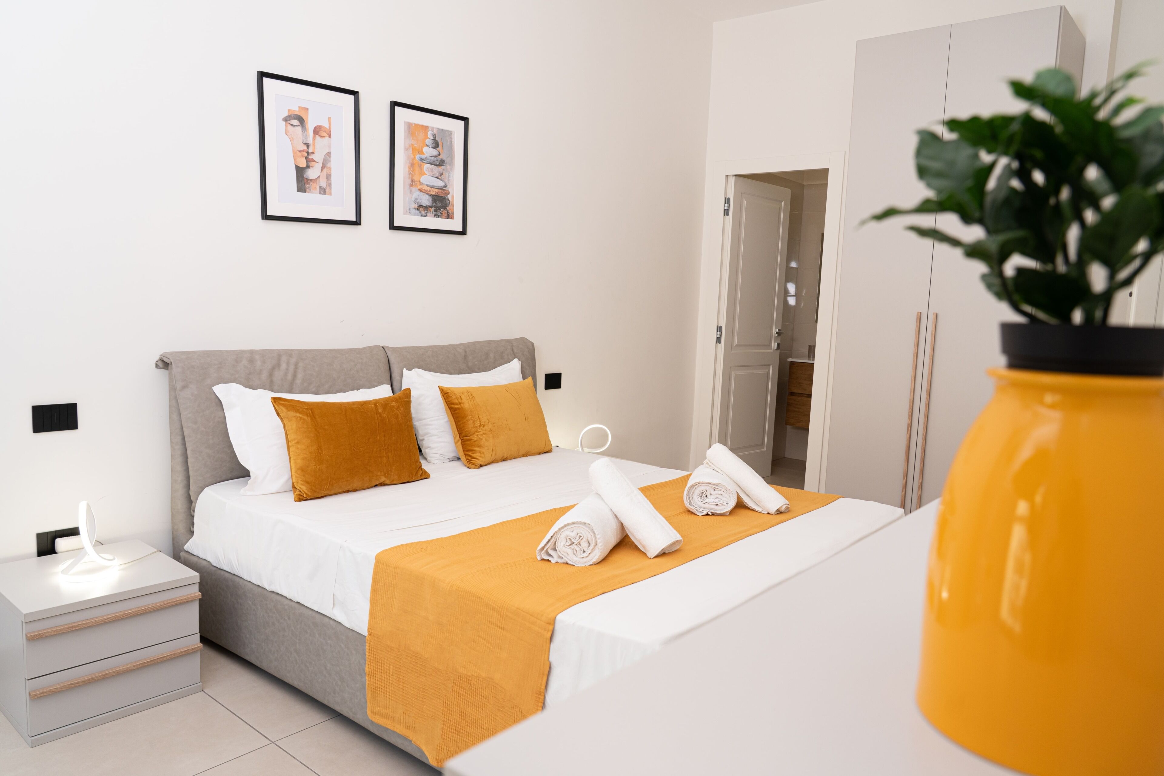 A modern bedroom with a bed, bedside tables, a nightstand, a potted plant, and two framed pictures on the wall.