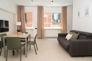 A modern living room with a sofa, a dining table with chairs, a flat-screen TV mounted on the wall, and large windows with curtains. - Via Niccolò Tommaseo 4 - Appartamento 1 (Naples)