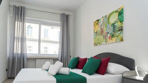 Una camera d'albergo con un letto, un comodino, una finestra con tende e un quadro appeso al muro.