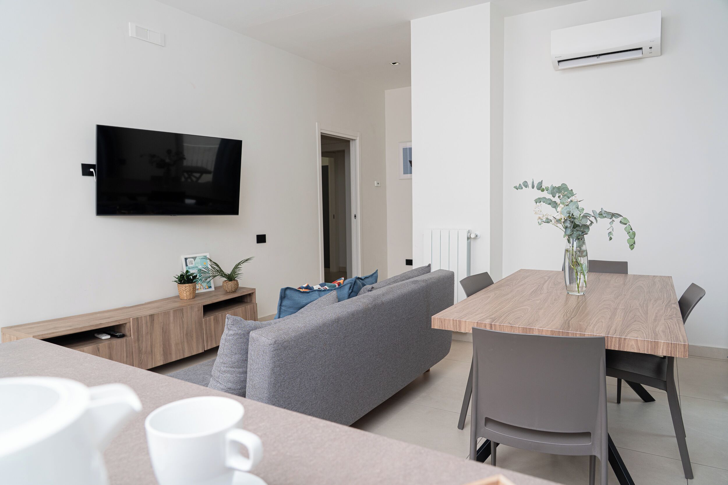A modern living room with a flat-screen TV, grey sofa, wooden dining table, and a small potted plant.