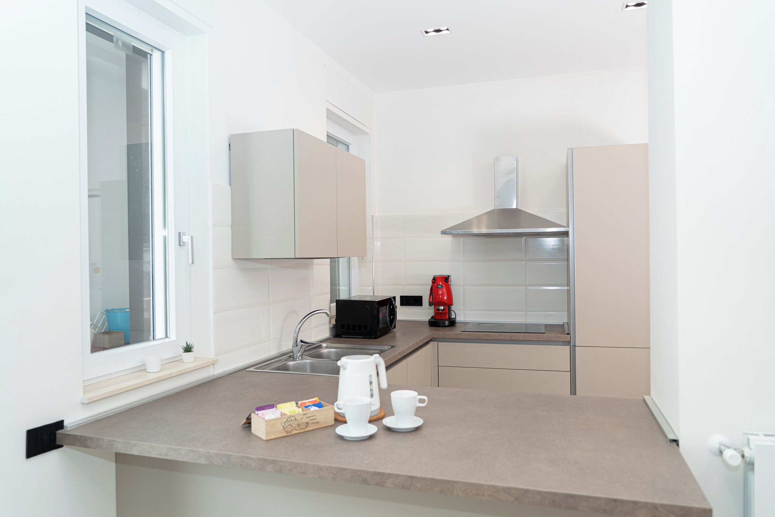 A compact kitchen with a window, a microwave, a red coffee maker, and a small coffee table with cups and a box of tea.