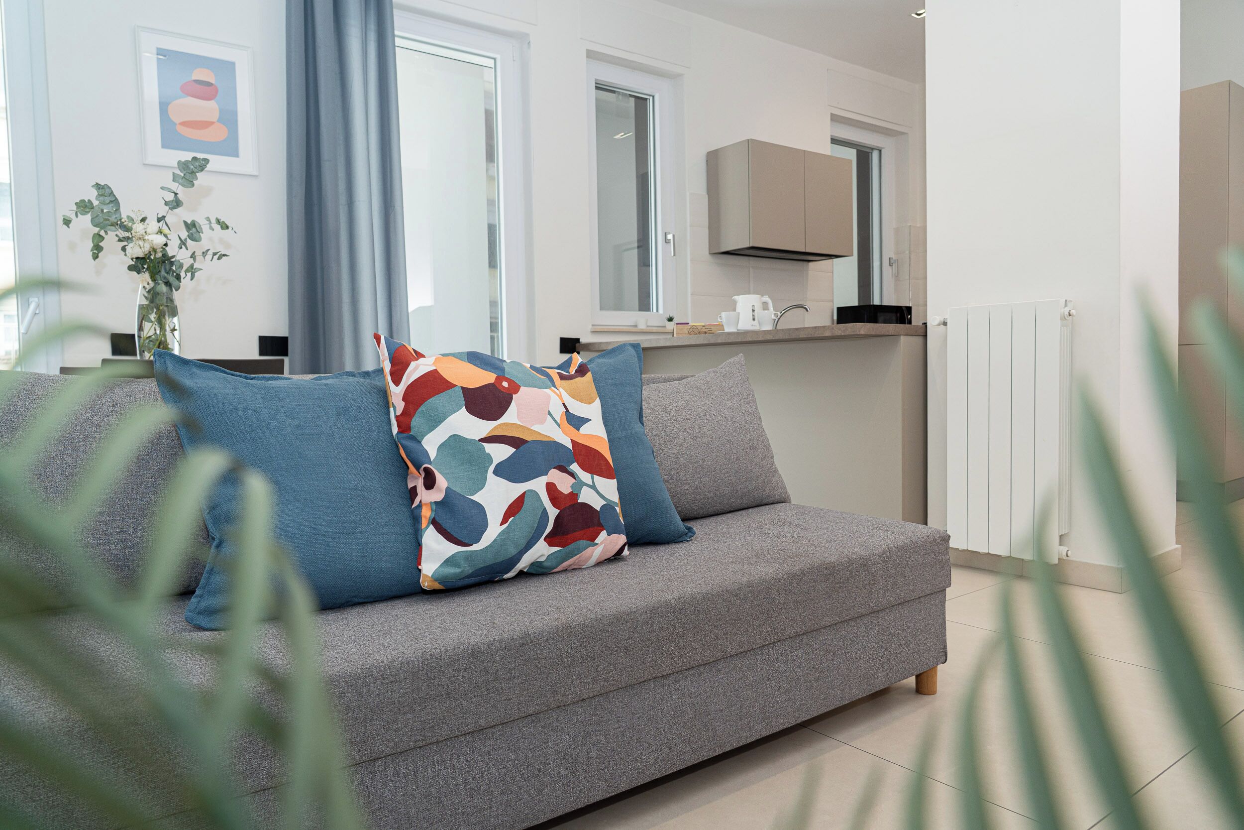 Un salon moderne avec un canapé gris, des coussins colorés, un vase de fleurs et une cuisine en arrière-plan.