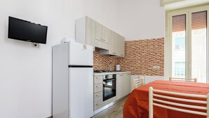 A compact kitchen with a refrigerator, oven, and microwave. There is a dining table with a red tablecloth and a chair. The kitchen has wooden cabinets and a tiled backsplash. - Via Morghen 63 - Primo Piano (Naples)