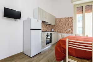 A compact kitchen with a refrigerator, oven, and microwave. There is a dining table with a red tablecloth and a chair. The kitchen has wooden cabinets and a tiled backsplash. - Via Morghen 63 - Primo Piano (Naples)