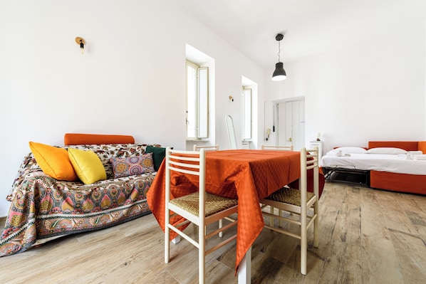 Una habitación con una mesa de comedor, un sofá con cojines de colores, una cama con ropa de cama naranja y suelo de madera.