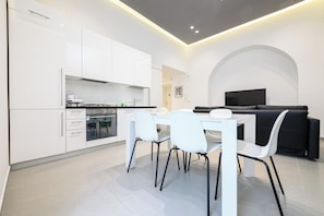 A modern kitchen with white cabinets, a dining table with chairs, and a black sofa in a living area with a TV. - Via Morghen 63 (Naples)